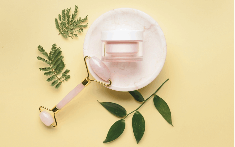 Face brightness cream is placed on white plate with jade roller beside and leaves in the background